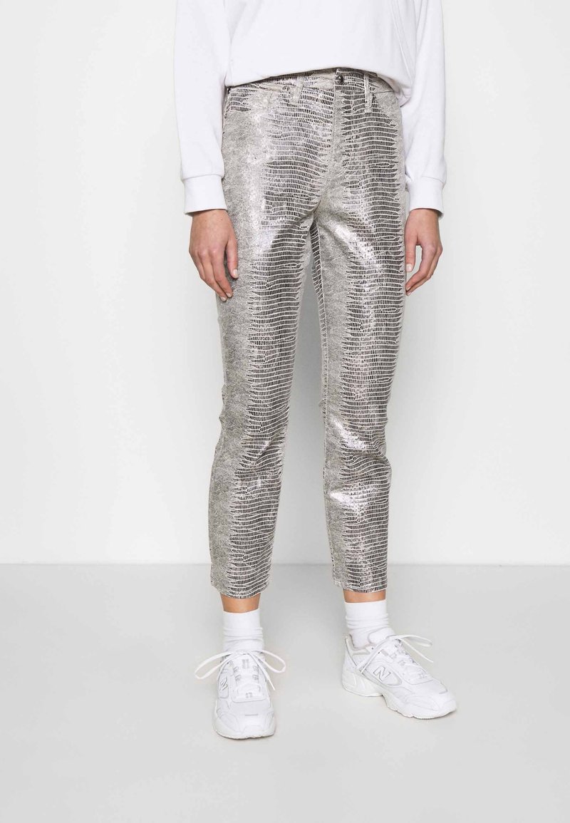 Pantalon argenté métallique avec un motif texturé inspiré des reptiles ; design fuselé, taille mi-haute, porté avec des baskets blanches et des chaussettes.