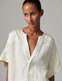 Μπλούζα σε κρεμ χρώμα με κοντά μανίκια, βαθύ V-λαιμόκοψη, απαλή υφή και διακριτική ραφή. Απλός, φαρδύς σχεδιασμός.