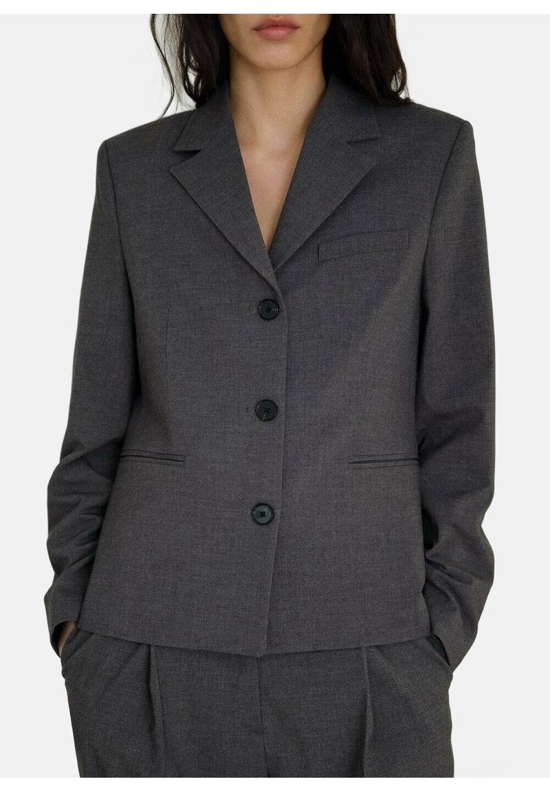 Femme portant un blazer gris foncé sur mesure avec trois boutons noirs et un pantalon assorti, mains dans les poches, sans accessoires visibles.