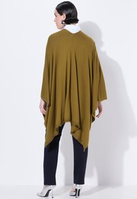 Grüner Poncho mit drapierter Gestaltung, fransigem Saum und subtilen Perforationen am Ausschnitt, getragen über navyfarbenen Hosen und silbernen Absätzen.