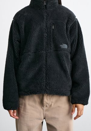 Person trägt eine schwarze Fleecejacke mit Reißverschluss und einer kleinen Reißverschlusstasche auf der Brust sowie beige Hosen vor einem schlichten Hintergrund.