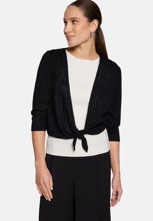 Frau trägt eine schwarze Strickjacke mit dreiviertellangen Ärmeln, die an der Taille gebunden ist, über einem weißen gerippten Oberteil und schwarzen Hosen, schaut zur Seite.