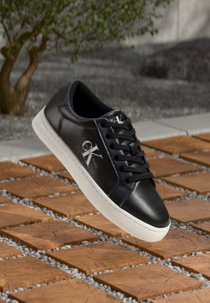 Black leather Calvin Klein sneaker with white sole, laces tied, displayed above wooden tiles and gravel outdoors near a tree.