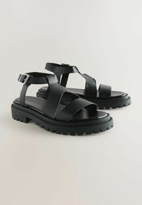 Svarta lädersandaler med justerbart ankle strap, öppen tå-design, kraftig texturerad sula och flera korsade remmar.