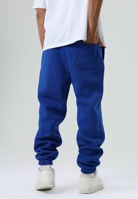 Blauwe katoenen joggingbroek met elastische boorden, een achterzak en een ontspannen pasvorm, gecombineerd met witte sportschoenen en zichtbare enkelsokken.