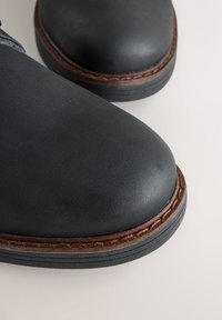 Ghete negre din piele pentru gleznă, cu vârfuri rotunjite, tălpi din cauciuc și cusături maro contrastante în jurul bazei. Textură netedă și design simplu.