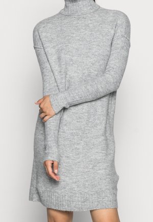 Kobieta ubrana w dzianinową sukienkę z długim rękawem i golfem w jasnoszarym kolorze, z rękami delikatnie skrzyżowanymi z przodu.