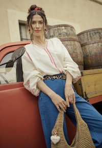 Donna appoggiata su un camioncino vintage rosso, che indossa una blusa bianca ricamata, jeans blu, una cintura leopardata, e tiene in mano una borsa intrecciata con un ciondolo a forma di cuore color crema.