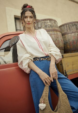 Kvinna lutad mot röd vintage-lastbil, iklädd en broderad vit blus, blåjeans, leopardbälte, hållande en vävd väska med en krämfärgad hjärtcharm.