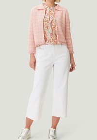 Giacca in tweed rosa chiaro con colletto e orlo sfrangiato, indossata sopra una blusa a fantasia. Abbinata a pantaloni bianchi a gamba larga e sneakers argentate.