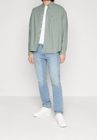 Chemise boutonnée vert clair portée sur un t-shirt blanc, associée à un jean bleu clair et des baskets blanches. La chemise possède une poche poitrine.
