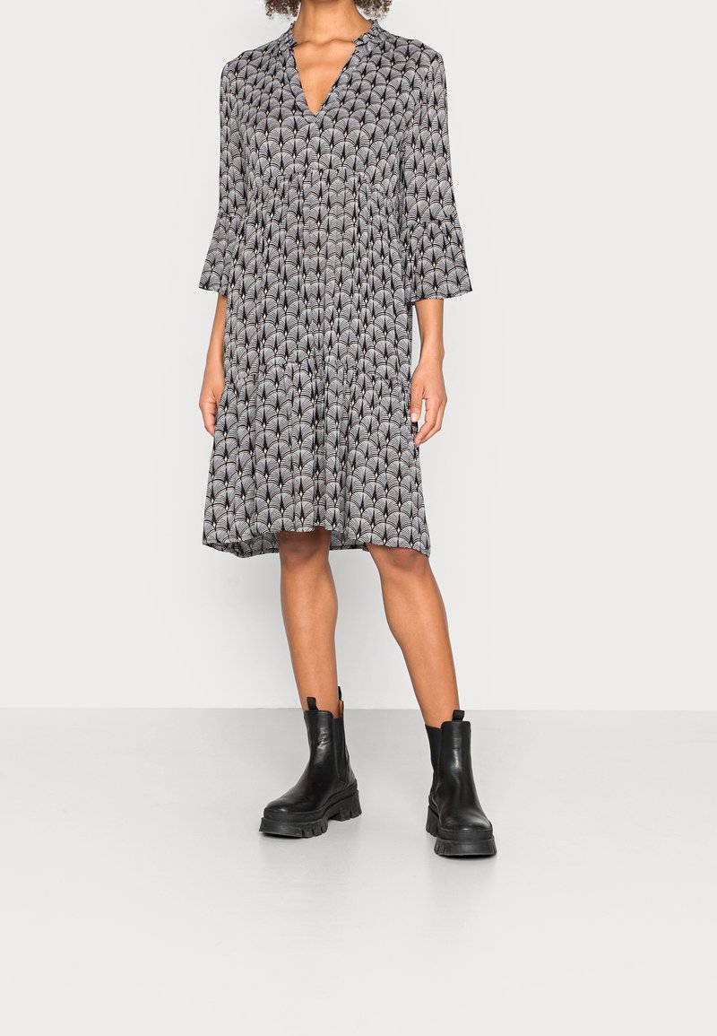 Robe noire et blanche à motifs avec un col en V, des manches cloche trois-quarts et une coupe ample, associée à des bottes noires épaisses.