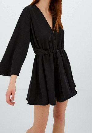 Woman wearing a short black wrap dress with wide long sleeves and a tied waist belt, standing against a plain light background.