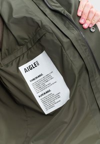 Olívová bunda s hladkou, matnou textúrou; obsahuje bielu nálepku s textom. Viditeľný zips a jemné švy.