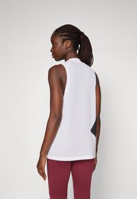 Canottiera atletica bianca in rete con un accento nero sul retro, caratterizzata da un taglio senza maniche e tessuto testurizzato, abbinata a leggings color mogano.