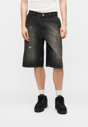 Személy fekete kifakult farmer rövidnadrágot visel kopott részletekkel, fehér zoknit és fekete tornacipőt, egyszerű háttér előtt áll.