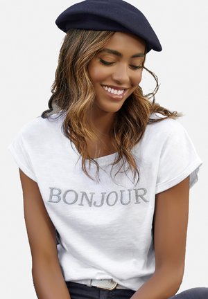 Weißes Baumwoll-T-Shirt mit dem Schriftzug "BONJOUR" in silberner Stickerei, mit aufgerollten Ärmeln, kombiniert mit einer navy-blauen Beretmütze. Glatte Textur und lässige Passform.
