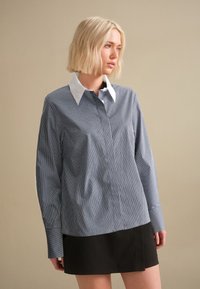 Striped blue and white long-sleeve shirt with a white collar, paired with a black skirt. Features buttoned sleeves and a relaxed fit.