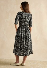 Schwarzes Blumenkleid mit weißen Blüten, 3/4-Ärmel, geraffte Taille, knielanger Rock, kombiniert mit hellen Slipschuhen.