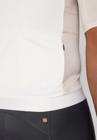 Lys beige cykeltrøje med en tætsiddende pasform, korte ærmer og en lynlåslomme på siden, parret med sorte shorts med logo-detalje.