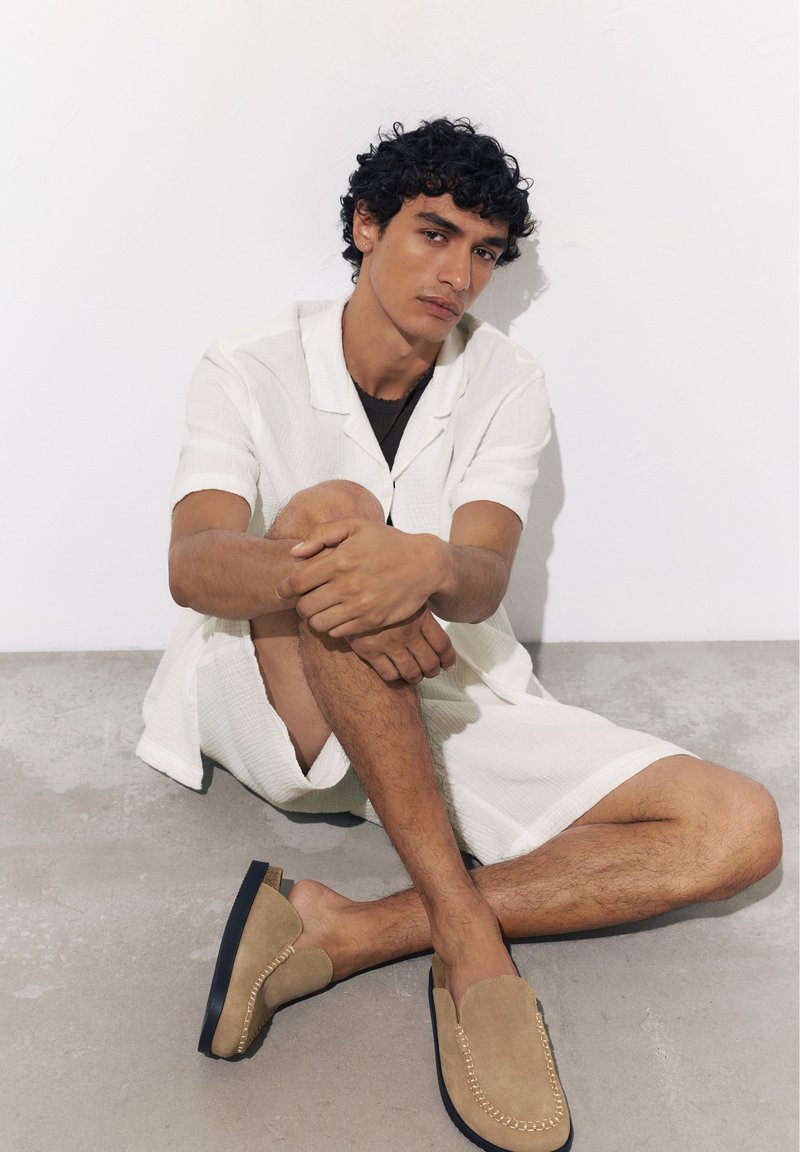 Jeune homme aux cheveux bouclés assis sur un sol gris, portant une chemise blanche à manches courtes, un short assorti et des chaussures beige à enfiler devant un mur blanc.