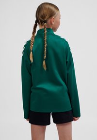 Green long-sleeve jacket with a stand collar, featuring white accents on the shoulders. Soft, smooth material, paired with black shorts.