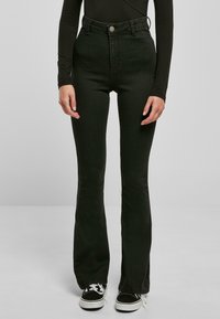 Mujer vistiendo jeans de campana negros de cintura alta, una blusa negra de manga larga y zapatillas en blanco y negro, de pie contra un fondo liso.