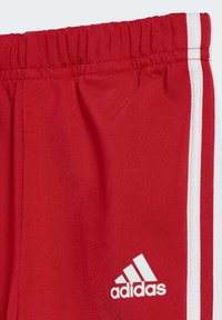 Pantalones deportivos rojos fabricados con una tela texturizada, con franjas laterales blancas y un logo de Adidas en blanco en la parte inferior izquierda.