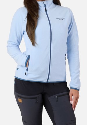 Kvinde iført lyseblå fleecejakke med lynlås med Swedemount-logo og sorte bukser med lynlåslommer og små svenske flagpatches.