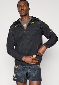 Μαύρο τζάκετ με φερμουάρ και γεωμετρικά σχέδια, με χρυσές λεπτομέρειες και κουκούλα. Φορεμένο πάνω από καφέ πουκάμισο και συνδυασμένο με μαύρο σορτς με σχέδια.