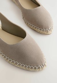 Espadrilles plates en daim taupe à bout pointu avec détails visibles de semelle en jute cousue à la main sur un fond clair.