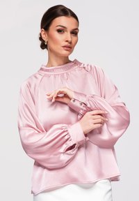 Femme posant dans un chemisier rose clair brillant à manches bouffantes et poignets boutonnés, associé à une jupe blanche, sur un fond uni.