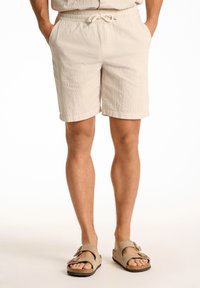 Beige shorts, jotka on valmistettu teksturoidusta kankaasta, elastisella vyötärönauhalla ja nyörillä. Yhdistettynä vaaleisiin, auki oleviin sandaaleihin.