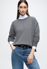 Γκρι, υπερμεγέθη μπλούζα με στρογγυλή λαιμόκοψη, φορεμένη πάνω από λευκό ζιβάγκο, συνδυασμένη με ανοιχτό μπλε τζιν και μαύρη ζώνη.