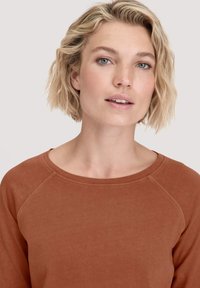 Blonde Frau mit kurzem, welligem Haar, die ein rostfarbiges Rundhalsoberteil trägt und mit dem Gesicht nach vorne vor einem hellen, neutralen Hintergrund steht.