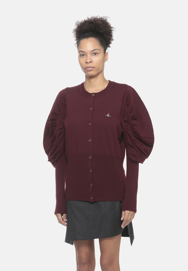 Donna che indossa un cardigan bordeaux con bottoni e maniche a sbuffo, insieme a una gonna grigia asimmetrica, in piedi contro uno sfondo neutro.