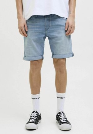 Homme portant un short en jean bleu clair retroussé, des chaussettes blanches avec le texte "Jack & Jones" et des baskets noires basses.