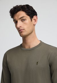 Olivgrön långärmad skjorta i mjukt material med rund halsringning och en liten broderad logga på bröstet. Slät textur.