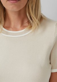 Kobieta nosząca beżowy krótki sweter z krótkim rękawem z białym wykończeniem przy kołnierzyku i krawędziach rękawów.