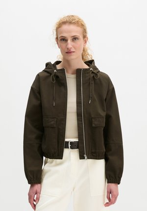 Femme aux cheveux blonds bouclés portant une veste à capuche vert foncé par-dessus un haut beige et un pantalon blanc avec une ceinture noire, debout devant un fond blanc.