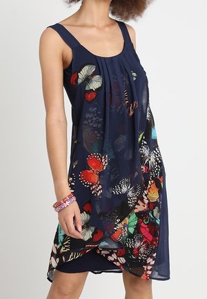 Ärmelloses dunkelblaues Kleid mit mehrfarbigem Schmetterlingsmuster, geschichteter Saum und geraffter Vorderseite, kombiniert mit pink gemusterten Armreifen am linken Handgelenk.