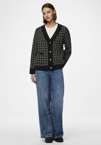 Kootud must kardigan valge ruudustiku mustriga, sügava V-kaelusega, nööpidega ja küljetaskutega; kombineeritud laia säärega siniste denimipedega.