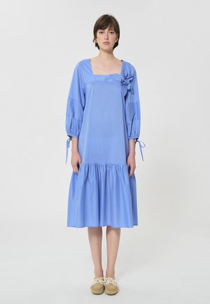 Mujer con vestido midi azul claro con escote cuadrado, mangas abullonadas con lazadas, falda fruncida, adorno floral y zapatos beige con cordones.