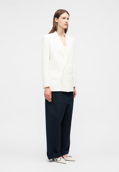 Femme portant un blazer blanc croisé, un pantalon large noir et des chaussures blanches à bouts pointus avec une boucle carrée, debout de profil sur un fond blanc.