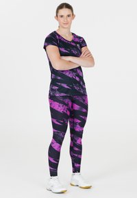 Tonårstjej som står med armarna i kors, klädd i svarta och lila mönstrade träningsleggings och en matchande kortärmad tröja med vita sneakers.