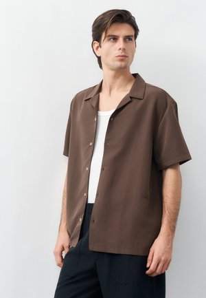 Νεαρός άνδρας με καφέ πουκάμισο με κοντά μανίκια και κουμπιά πάνω από λευκό φανελάκι, μαύρο παντελόνι, κοιτάζει ελαφρώς προς τα αριστερά του, μπροστά σε απλό φωτεινό φόντο.