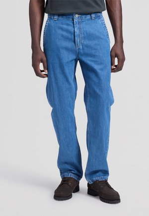 Uomo che indossa jeans azzurri a gamba dritta, scarpe stringate marrone scuro e una camicia scura, in piedi davanti a uno sfondo chiaro uniforme.