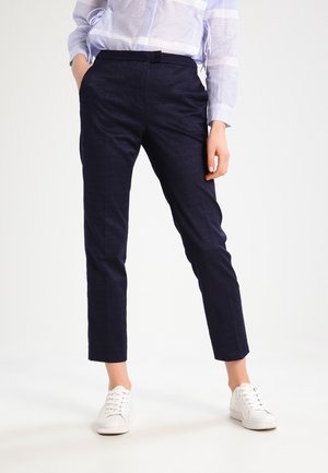 Person trägt dunkelblaue, maßgeschneiderte Hose, hell gestreiftes Langarmhemd, weiße Sneakers, steht mit einer Hand in der Tasche.