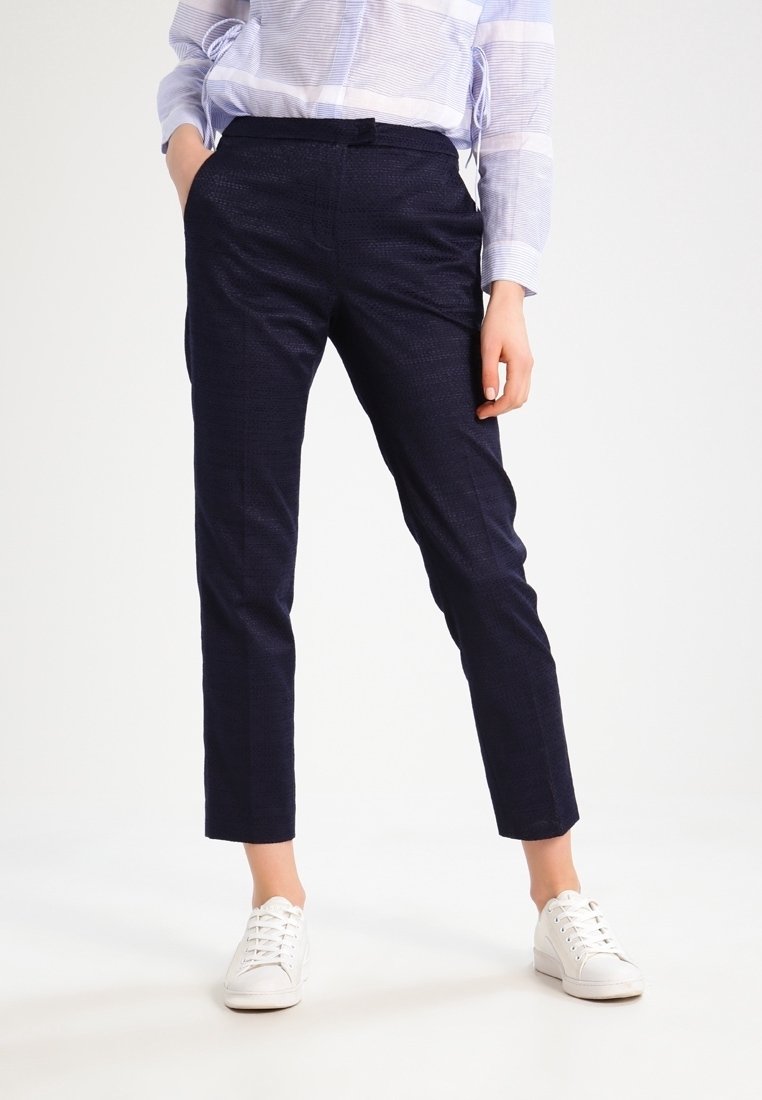 Personne portant un pantalon bleu marine foncé sur mesure, une chemise légère à rayures manches longues, des baskets blanches, debout avec une main dans la poche.