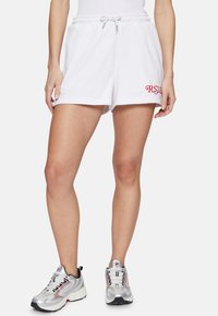 Pantalones cortos de algodón blanco con una cinturilla elástica y cordón, con bordado rojo de "RSVP" en el lado izquierdo. Combinados con zapatillas deportivas plateadas.